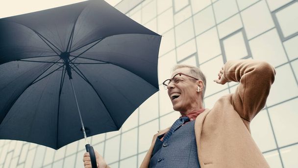 Low angle view of positive businessman in earphone holding umbrella on urban street  - Foto, Bild