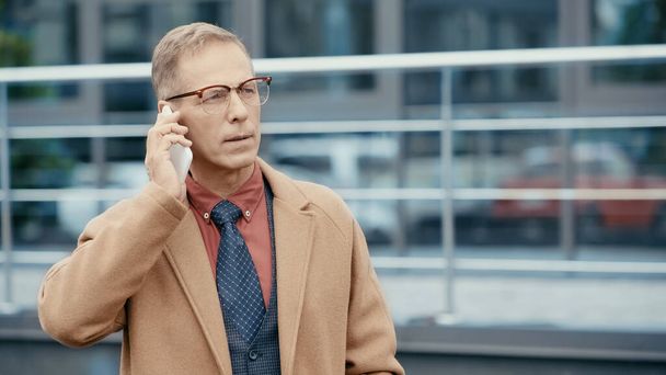 Mature businessman in coat talking on mobile phone on urban street  - Photo, Image