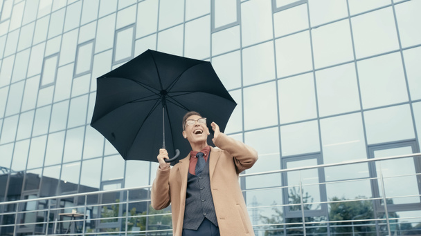 Feliz hombre de negocios con abrigo y auriculares sosteniendo paraguas en la calle urbana  - Foto, Imagen