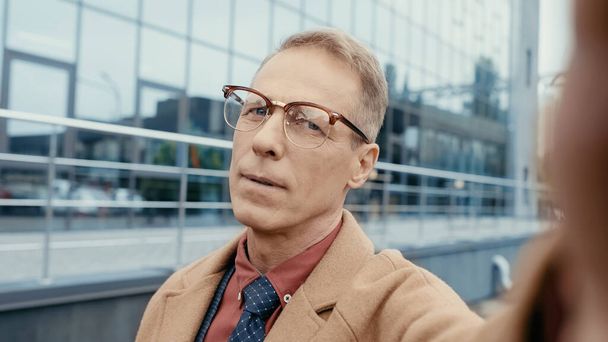 Middle aged businessman in eyeglasses and coat looking at camera outdoors  - Foto, immagini