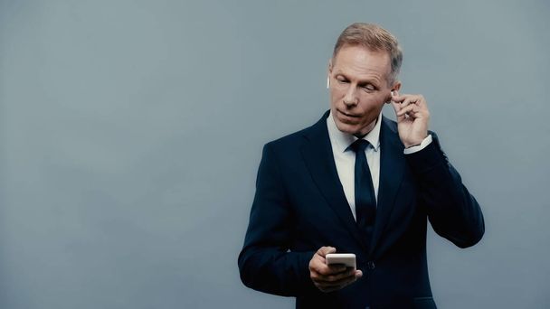 Mature businessman in formal wear using smartphone and earphones isolated on grey  - Фото, изображение