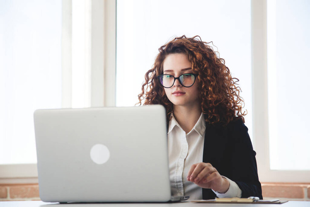 Portret of a successful young curly red-haired woman in a business suit and glasses works in an office on a laptop - 写真・画像