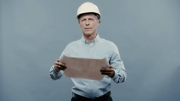 Middle aged engineer in hardhat holding blueprint and looking at camera isolated on grey  - Foto, Imagem
