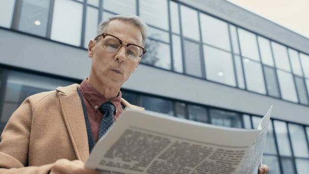 Businessman in coat reading newspaper near blurred building outdoors  - Foto, afbeelding