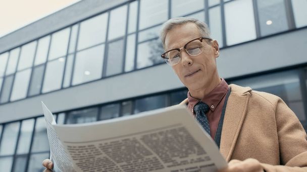 Low angle view of smiling businessman in coat reading newspaper outdoors  - 写真・画像