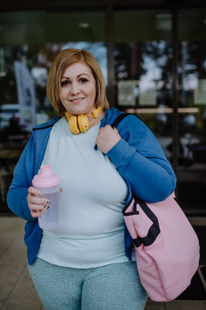 Mujer feliz con sobrepeso en ropa deportiva mirando a la cámara al aire libre frente al gimnasio - Foto, Imagen