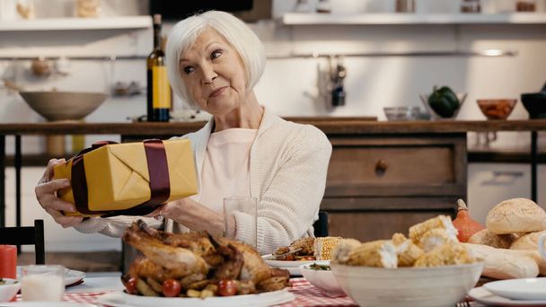 pleased senior woman holding present near served thanksgiving dinner - Фото, изображение