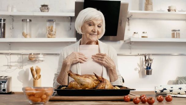 χαρούμενη γυναίκα που κοιτάζει νόστιμη γαλοπούλα κοντά σε φρέσκα ντοματίνια και καρότα στην κουζίνα - Φωτογραφία, εικόνα