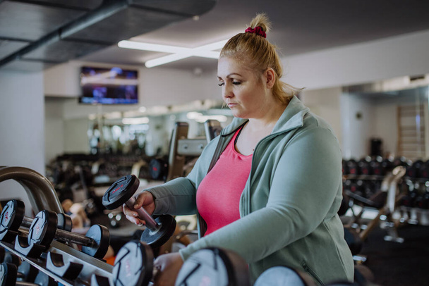 Plus size woman training taking dumbbells indoors in gym - Foto, afbeelding