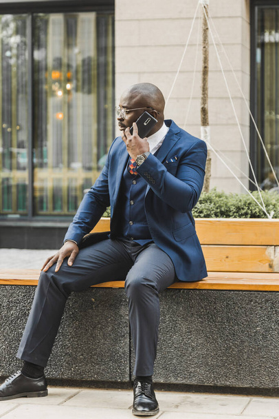 Ritratto di un uomo d'affari afroamericano nero in giacca e cravatta seduto su una panchina in un parco cittadino all'aperto. - Foto, immagini