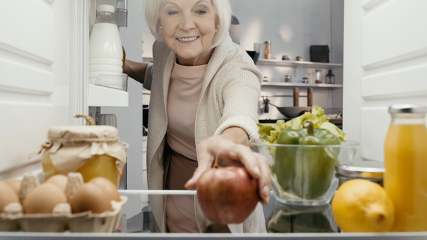 happy senior woman taking fresh apple from fridge with fresh vegetables, fruits, drinks and eggs - Fotó, kép