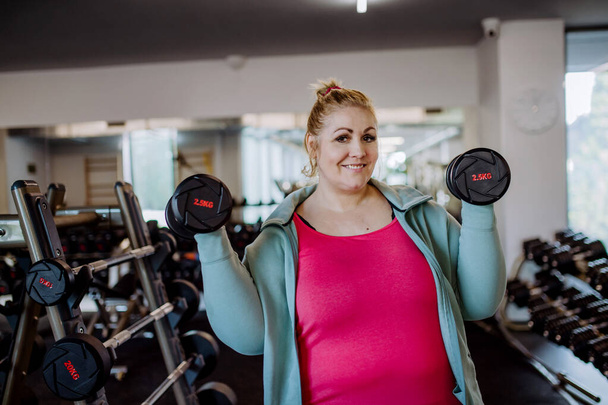Plus size woman training and lifting dumbbells indoors in gym, looking at camera - Fotografie, Obrázek