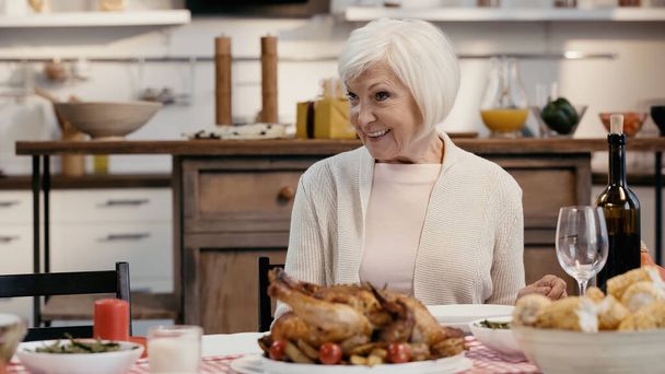 happy senior woman sitting near roasted turkey and red wine during thanksgiving dinner - Фото, изображение