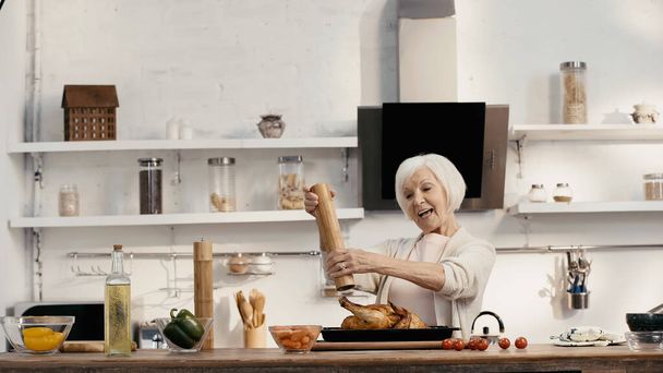 χαρούμενη ηλικιωμένη γυναίκα καρυκεύει ψητή γαλοπούλα κοντά σε φρέσκα λαχανικά και λάδι στην κουζίνα - Φωτογραφία, εικόνα