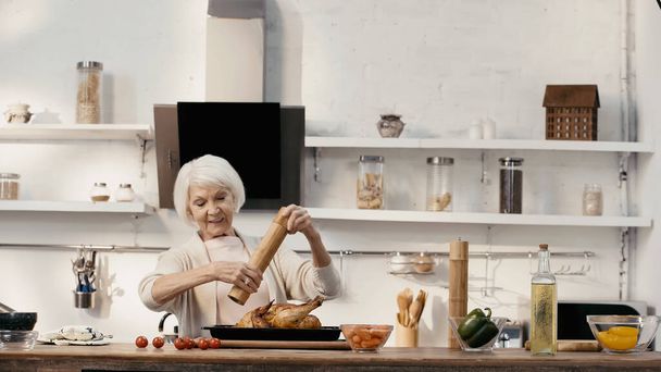 усміхнена старша жінка приправляє смажену індичку біля свіжих овочів та олії на столі
 - Фото, зображення