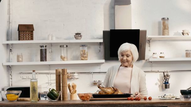 pleased senior woman looking at delicious turkey near fresh vegetables and spice mills in kitchen - Foto, Imagen