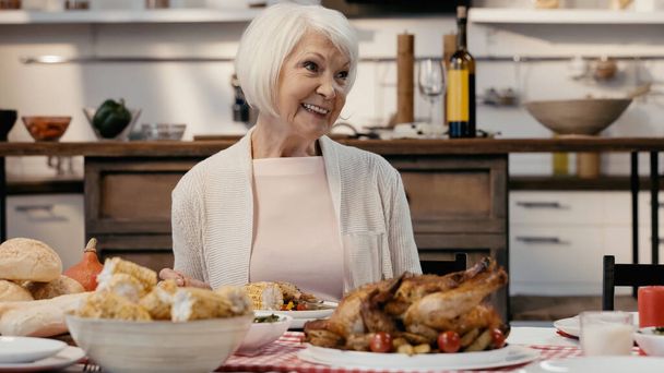 Ευτυχισμένη ηλικιωμένη γυναίκα χαμογελά κοντά στο δείπνο των Ευχαριστιών σερβίρεται στην κουζίνα - Φωτογραφία, εικόνα