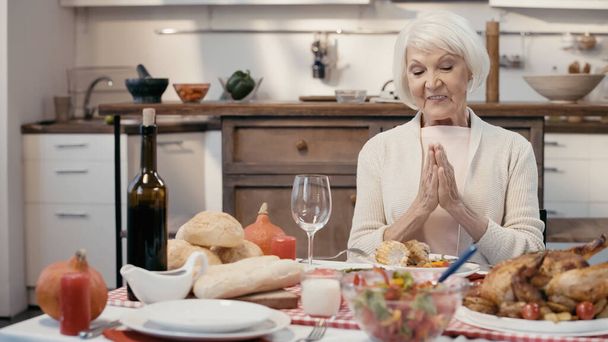 sonriente anciana rezando antes de la cena de acción de gracias cerca de pavo asado con verduras, vino tinto y bollos - Foto, Imagen