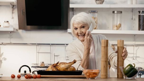 alegre mujer mayor hablando en el teléfono celular cerca de pavo asado, molinos de especias y verduras frescas - Foto, Imagen