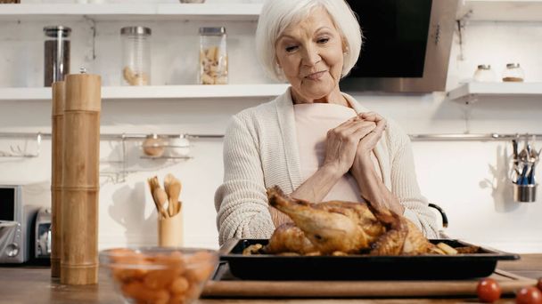pleased senior woman looking at delicious turkey near spice mills in kitchen - Фото, изображение