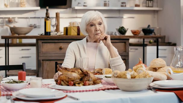 lonely and thoughtful senior woman sitting near thanksgiving dinner served on table in kitchen - Photo, Image