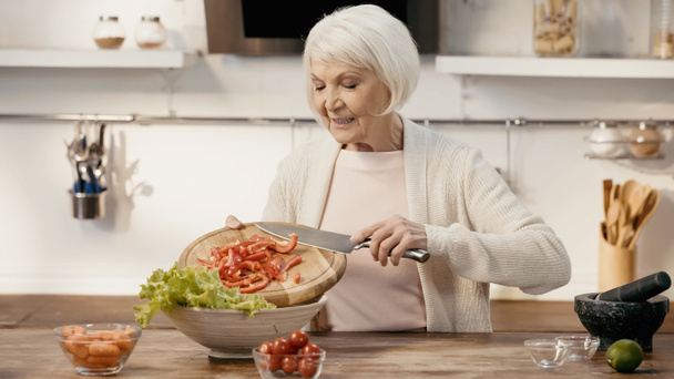 mujer mayor sonriente agregando pimiento en rodajas a la lechuga mientras prepara ensalada de verduras - Foto, imagen