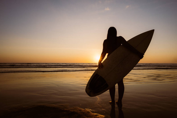 Gün batımında kumlu sahilde sörf tahtası olan güzel seksi sörfçü kadın silueti. Su sporları. Sörf sağlıklı aktif yaşam tarzıdır. Yaz tatili. - Fotoğraf, Görsel