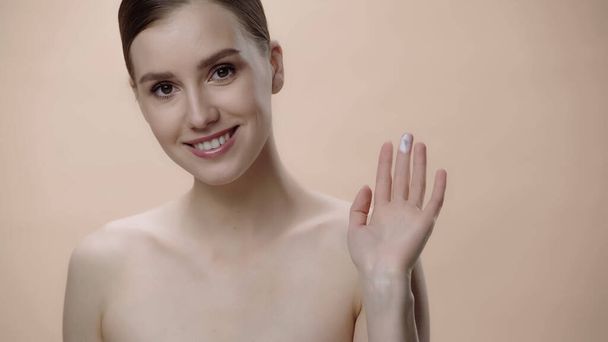happy young woman with bare shoulders showing fingers with face cream isolated on beige  - 写真・画像