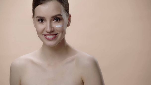 happy young woman with bare shoulders and cream on face isolated on beige  - Foto, afbeelding