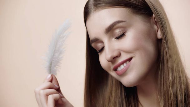 happy young woman with closed eyes holding feather isolated on beige  - 写真・画像