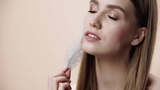 young woman with closed eyes holding feather near face isolated on beige  - Foto, Imagen