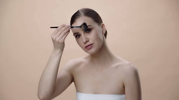 young woman with bare shoulders applying face powder with cosmetic brush isolated on beige - Foto, afbeelding