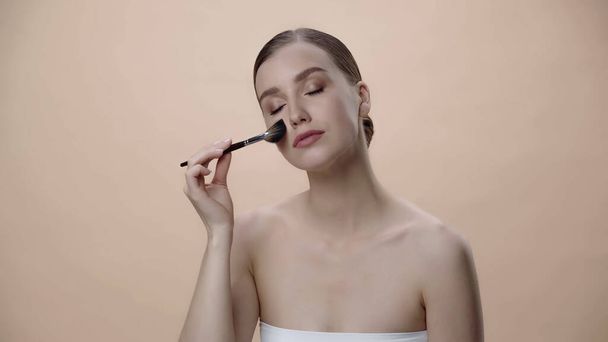 young woman with bare shoulders and closed eyes applying face powder isolated on beige - 写真・画像