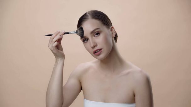 young woman applying face powder while holding cosmetic brush isolated on beige - Foto, Bild