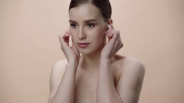 young woman with bare shoulders adjusting earphones isolated on beige  - Fotó, kép