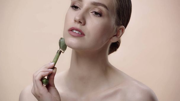 young woman with bare shoulders massaging face with jade roller isolated on beige  - Fotografie, Obrázek