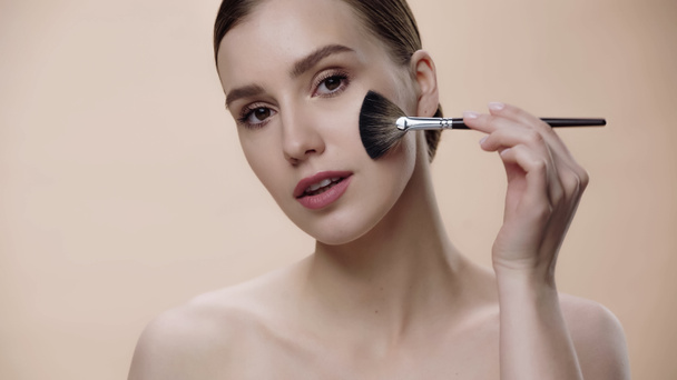 young woman with bare shoulders applying face powder on cheek isolated on beige - Foto, immagini