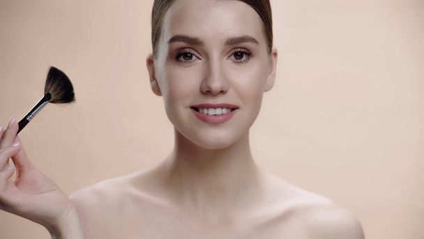 joyful woman with bare shoulders holding cosmetic brush isolated on beige - Photo, image