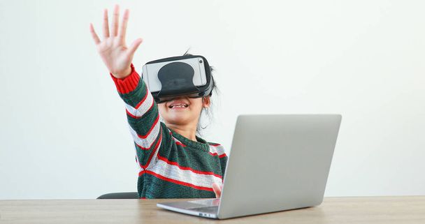 La niña que lleva VR sentada en el escritorio en las gafas de casa virtual Global Internet conexión metaverse. Futuros niños en ropa blanca con auriculares VR para un juego. concepto tecnología avanzada.  - Foto, imagen