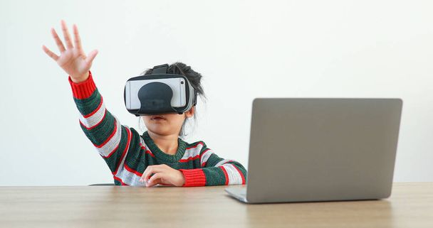 Pieni tyttö, jolla on VR, istuu pöydän ääressä kovalaseissa virtuaalisessa Global Internet-yhteydessämetaverse. Tulevat lapset valkoisissa vaatteissa yllään VR kuulokkeet peliin. käsite kehittynyttä teknologiaa.  - Valokuva, kuva