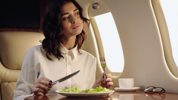 Pleased businesswoman holding cutlery near fresh salad and cup in private jet  - Fotografie, Obrázek