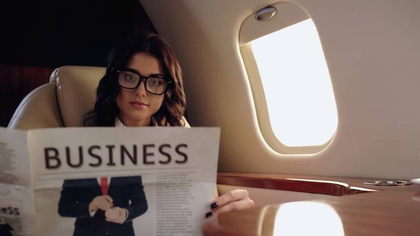 Νεαρή επιχειρηματίας με γυαλιά ηλίου που διαβάζει ειδήσεις σε ιδιωτικό αεροπλάνο  - Φωτογραφία, εικόνα