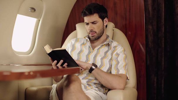 Bearded man sticking out tongue while reading book in private plane  - Foto, Imagem