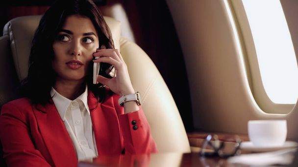 Businesswoman talking on cellphone near blurred cup in private plane  - Foto, afbeelding