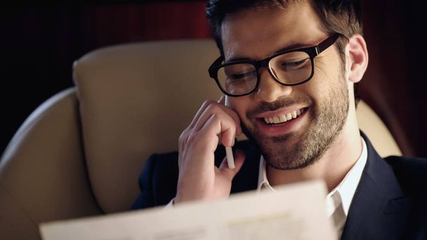 Hombre de negocios sonriente hablando por celular y leyendo el periódico en jet privado  - Foto, imagen