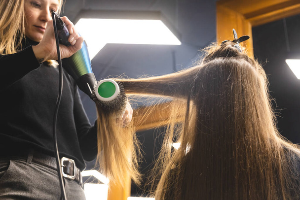 Master woman hairdresser dries the girls hair with a hairdryer and combs after washing in the beauty salon - 写真・画像