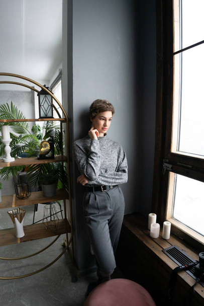 young beautiful woman is standing near the window in a sweater - Фото, зображення