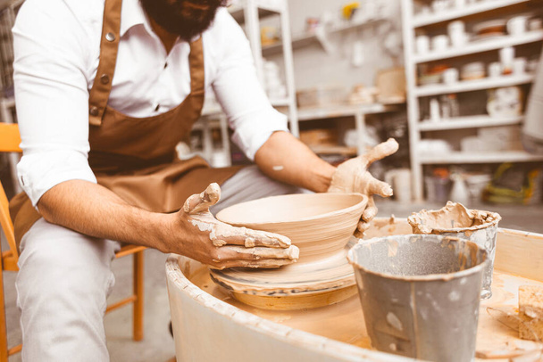 A young male potter is engaged in craft in his workshop on a potters wheel and makes a clay product - Zdjęcie, obraz