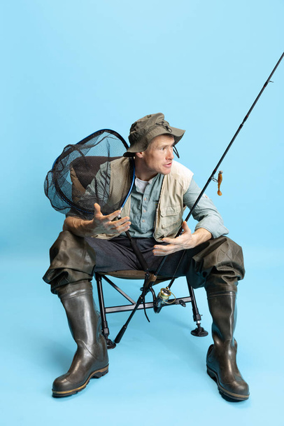 Portrait of young man, fisherman with fishing rod, spinning and equipment sitting isolated over blue studio background - Фото, зображення
