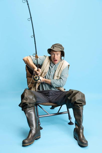 Retrato de joven, pescador con caña de pescar, spinning y equipo sentado aislado sobre fondo azul estudio - Foto, imagen